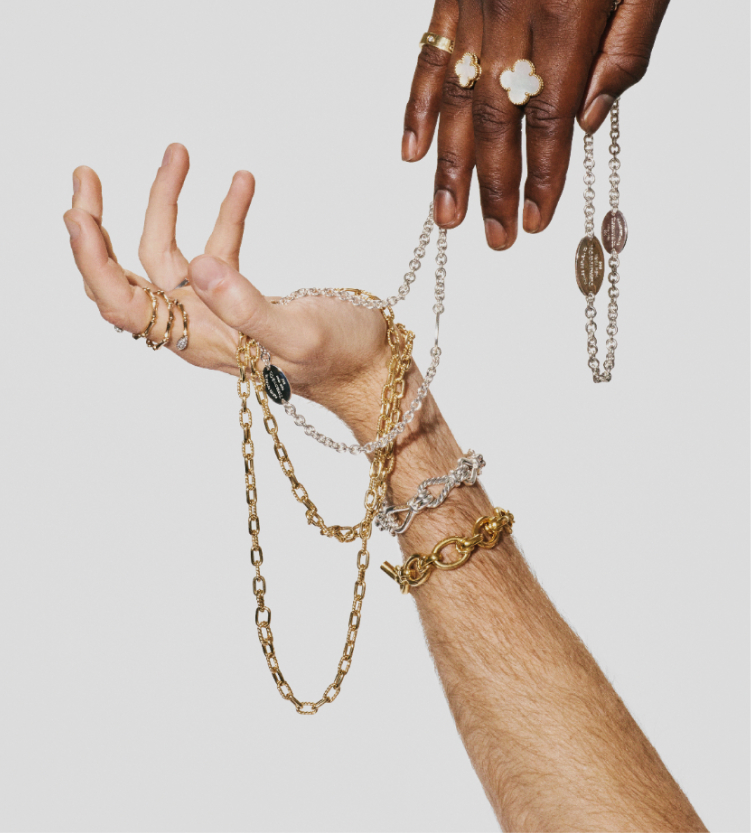 Two posed hands wearing rings with bracelets and necklaces strewn about on light gray background