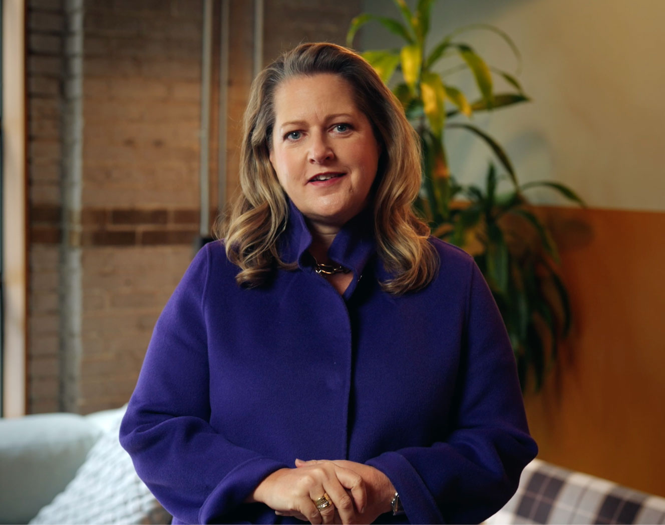 Featured video of a blonde woman in a purple coat, speaking before a brick wall and plants. Play button overlay visible.