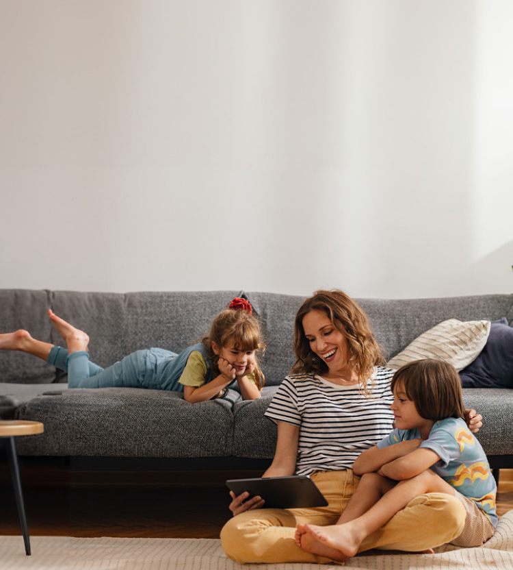 A mother sitting on the floor with her son and her daughter on the couch behind her. They’re all smiling and looking down at a tablet.
