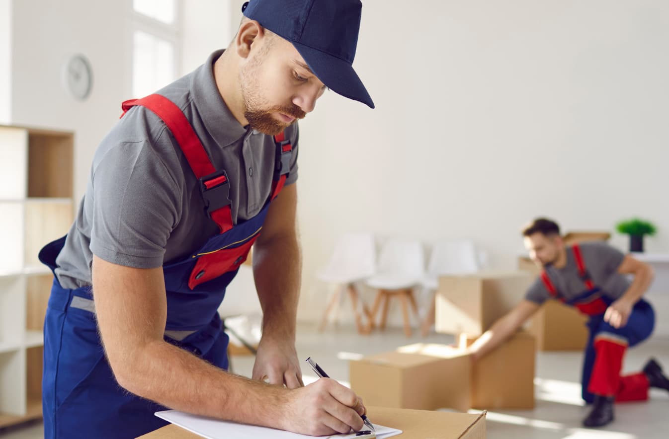  Man in a gray polo shirt, blue cap and overalls, bent over a box, writing on a piece of paper with another person reaching down for a cardboard box on the floor in the background.