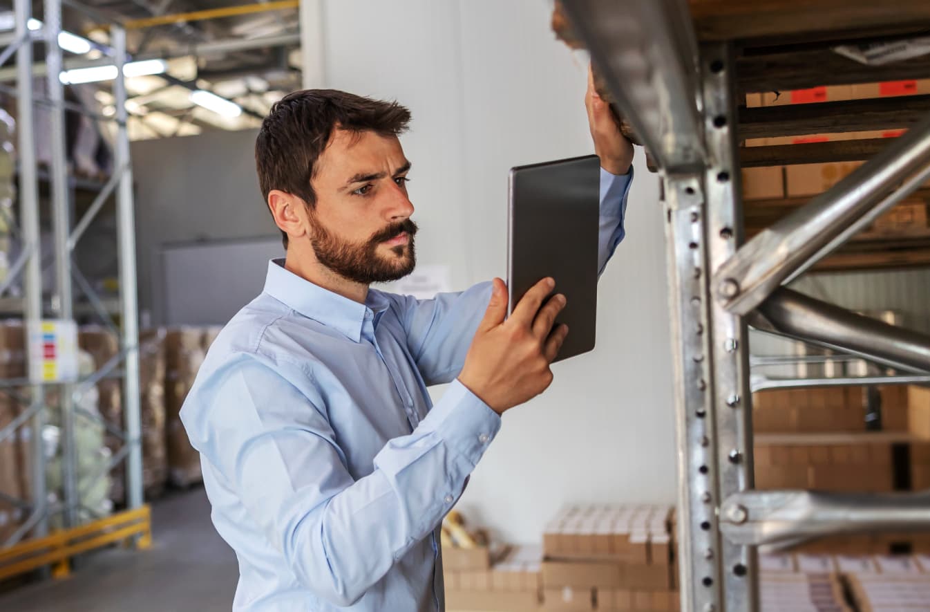 Man in a light-blue button-up standing in front of metal racks and surrounded by boxes, concentrating on the tablet in his hand.