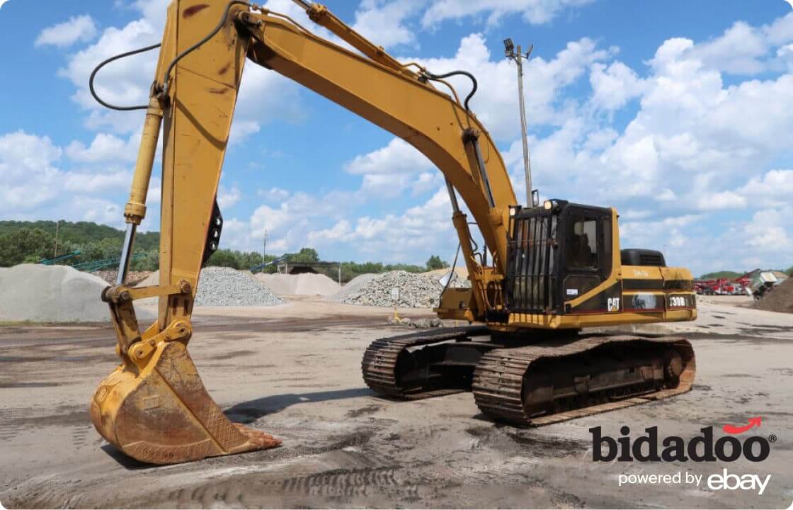  A yellow Caterpillar excavator is parked outside on a clear day with its scoop resting on the flat dirt ground. In the background, there are many mounds of dirt and gravel and a green forest strip stretches along the horizon. 