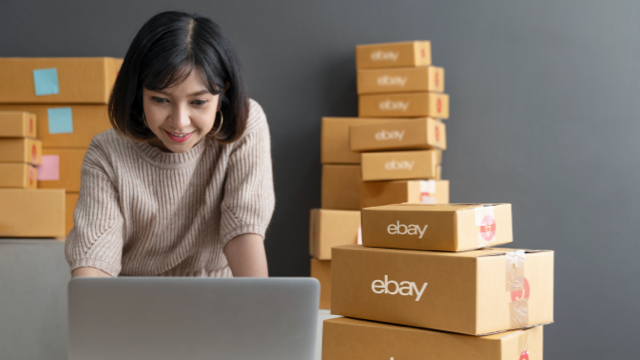 A smiling woman standing at a table, looking at her laptop, with boxes around her.