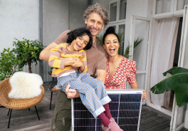 Eine dreiköpfige Familie lacht in die Kamera, der Vater hält das Kind im Arm, die Mutter ein Solarmodul vor sich.