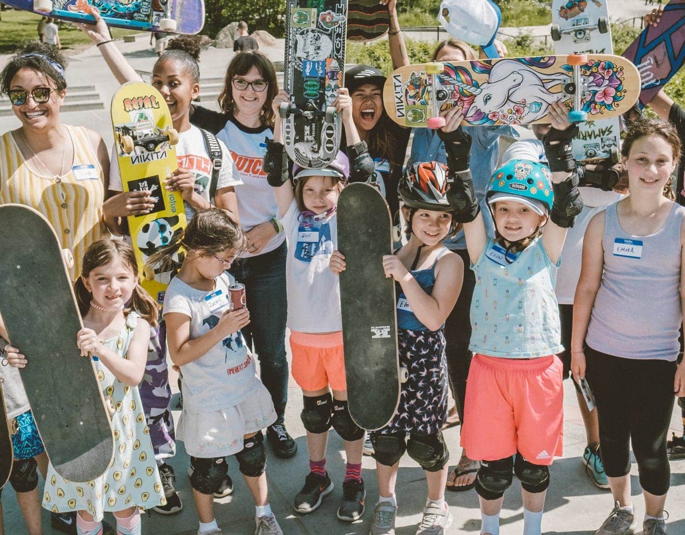 A group of kids of diverse ethnicities smiling at the camera, holding up their skateboards. Some of them are wearing helmets and knee and elbow pads.