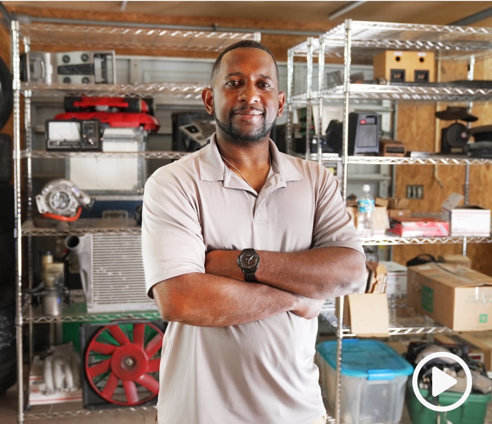 Seller James Patrick smiling at the camera with metal shelves full of car gear and equipment behind him.