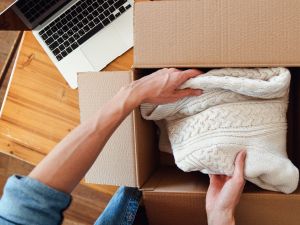 A close-up of a man packing a knitted, cream jumper into a cardboard box.