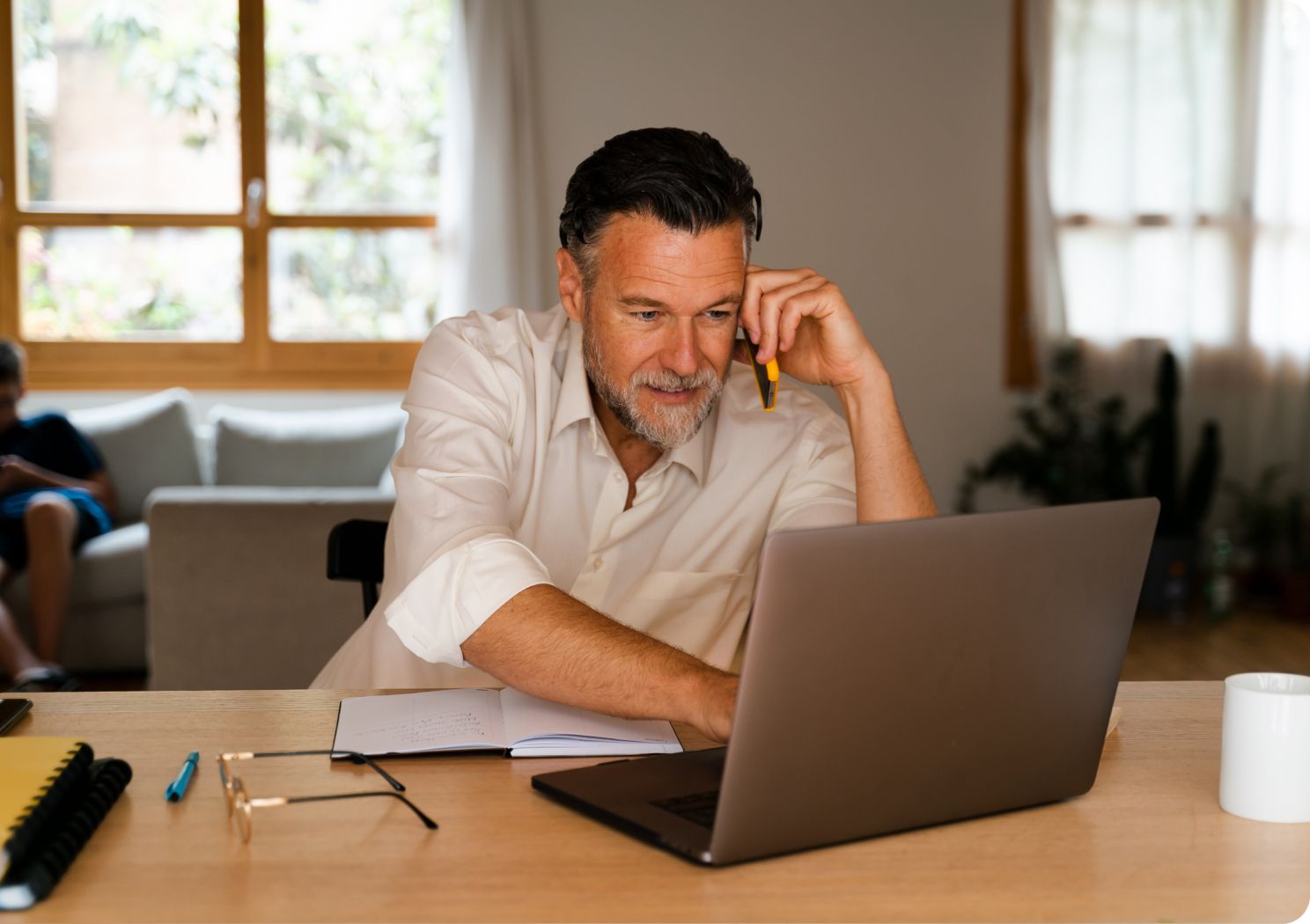 A man in a white shirt sits at a desk, talking on the phone whilst working on his laptop, with a notebook and glasses besides him.