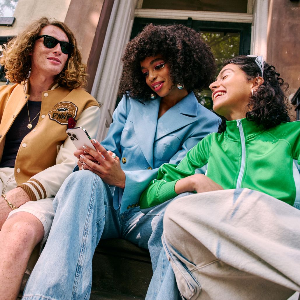 Three friends sit on a porch smiling while one looks at their smartphone.