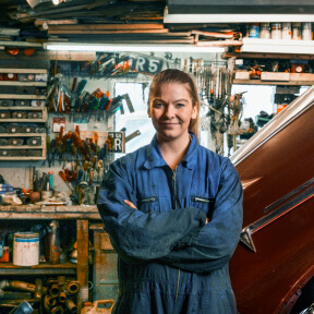 A woman in overalls smiling in a garage full of tools and a car.