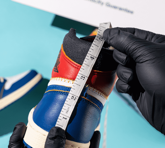 A pair of gloved hands measuring the length of the back of a sneaker. The other sneaker in the back is also in-frame lying on a light blue background.