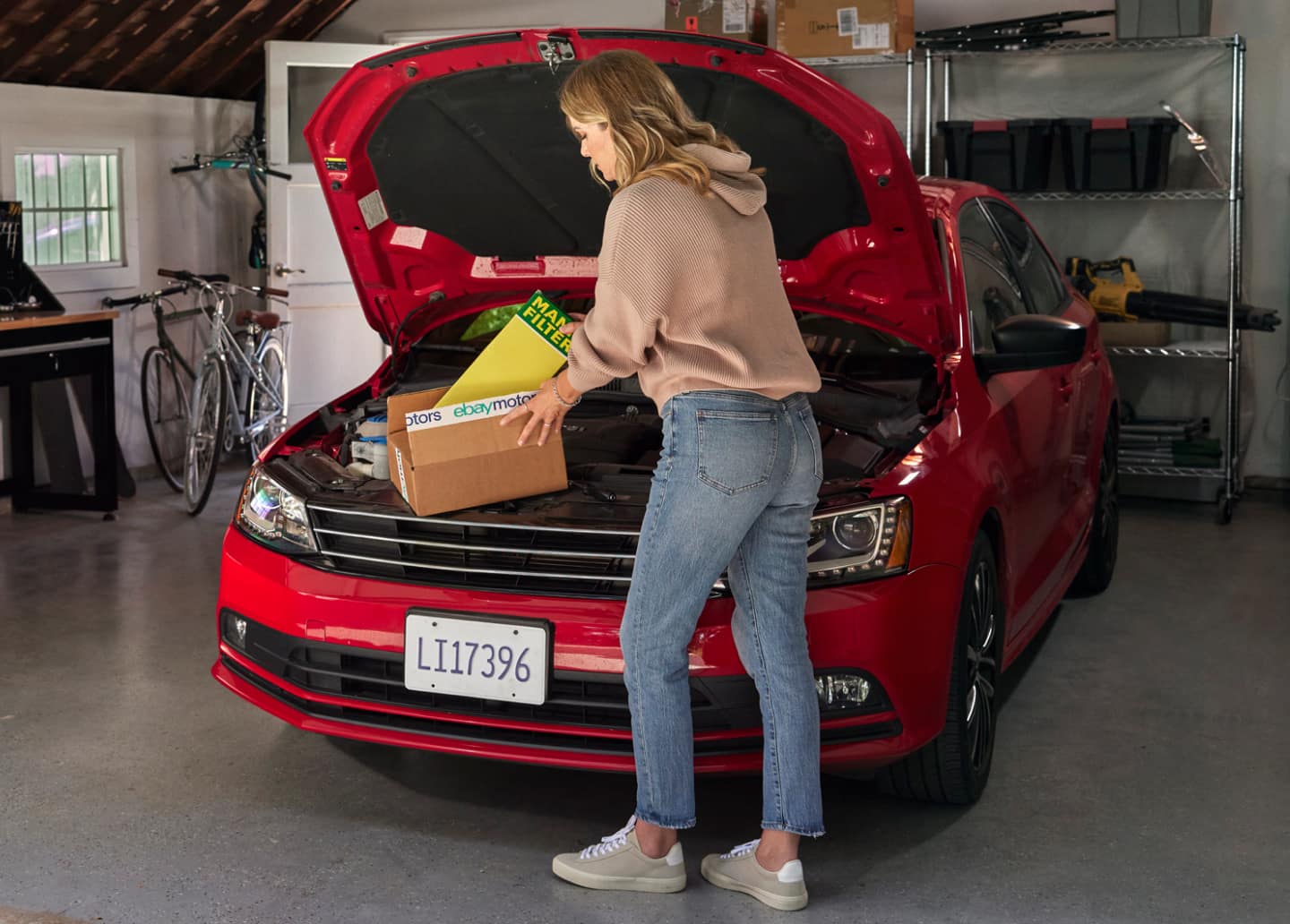 A woman in blue jeans and a salmon hoodie, standing in her garage, at the open hood of her red sedan, removes a yellow air filter out of an eBay Motors package with her sleeves rolled up. The garage floor is a gray polished concrete and in the background are three bicycles behind an open door and a metal rack with a toolbox, a tripod, and a leaf-blower.