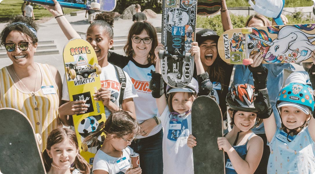 Photograph of young girls holding up their skateboards as they smile into the camera.