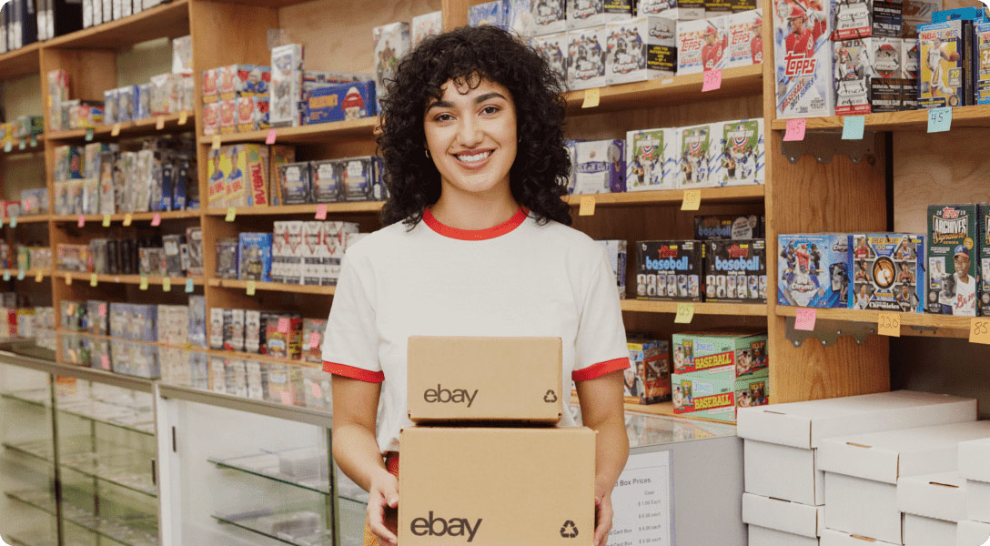 Photograph of a woman smiling into the camera as she stands infront of shelves full of inventory, while holding eBay shipping boxes.