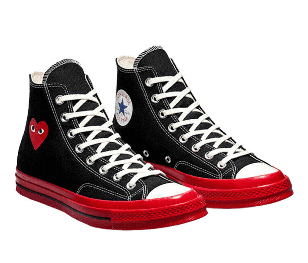 A pair of white and black COMME des GARÇONS Converse Chuck 70s. One side of the shoe has the Converse All Star logo, while the other side has the red COMME des GARÇONS heart logo. The entire sole of the shoes are red.