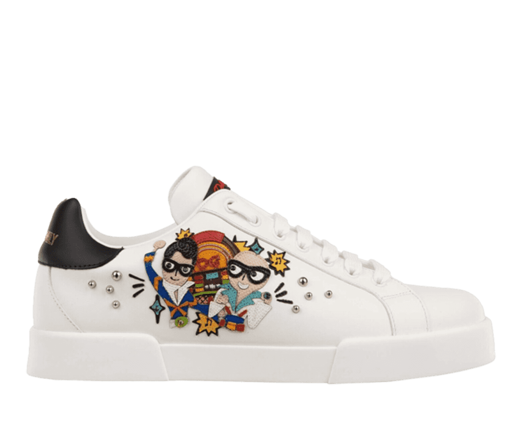A side shot of a white Dolce & Gabana sneaker. On the side of the shoe is a cartoon version of two men from the waist up. One is bald, the other has a pampadour hairstyle, and both are wearing colourful tuxedos and domino masks. A Dolce & Gabana branded jukebox is behind them, as well as musical notes and several beads.