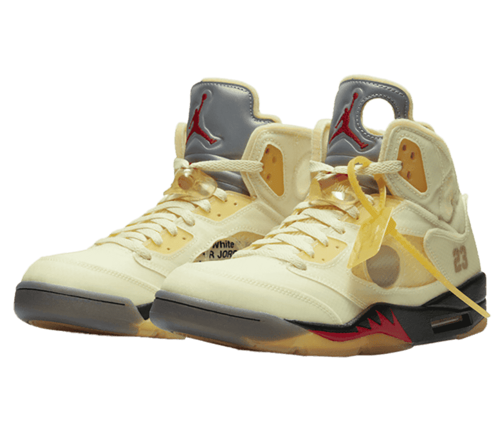 An yellowish off-white pair of AJ5 Retro sneakers in suede and plastic with translucent detailing. The shoes have lace cages, a beige plastic zip-tie tag on the toplace, and black midsoles with a red spiked pattern in the middle.