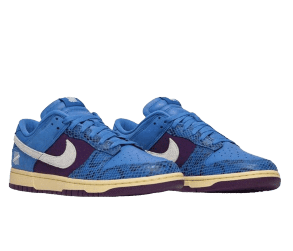 A front, angled view of the UNDEFEATED x Nike “Dunk Vs. AF-1”. The shoe has bold blue laces, vamp, collar, tongue, and inner lining. The mid-panel and sole are in a royal purple canvas and midsole in an off white, yellowish stain. The shoes are topped off with a white, nubuck Nike swoosh and the UNDEFEATED brand’s five-strike emblem on the tongue label and heel, also in white.