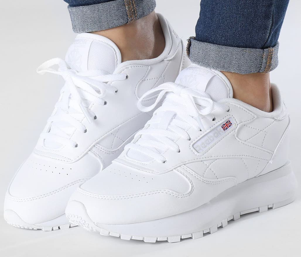 A close-up of a person's feet wearing an all-white pair of Reebok Classic sneakers and blue jeans with rolled-up cuffs.
