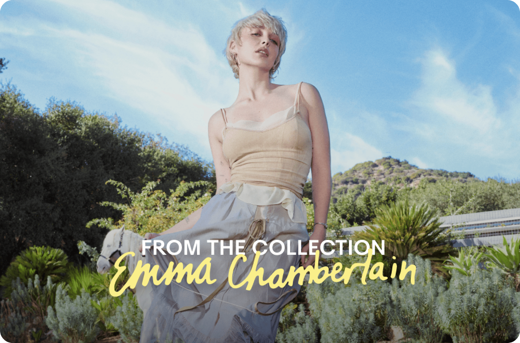 Emma Chamberlain posing outdoors in front of greenery and blue sky, wearing a beige top and flowing skirt, with text reading "From the Collection Emma Chamberlain."